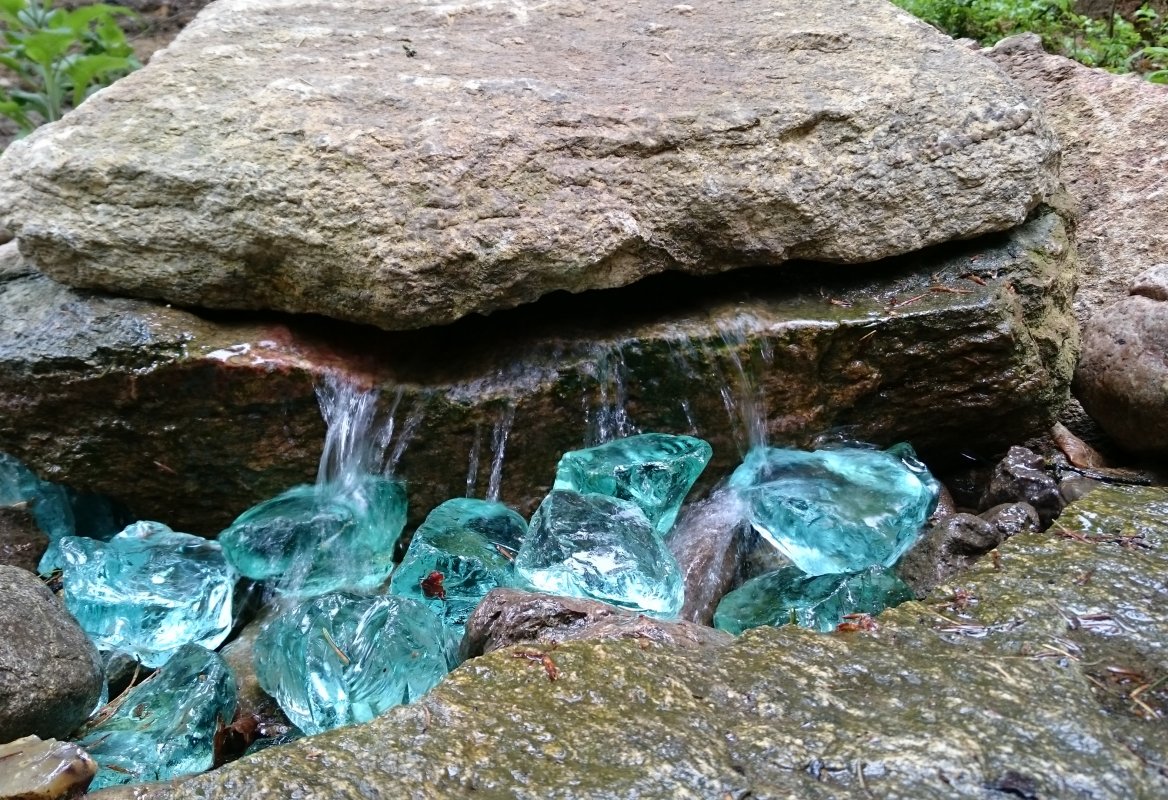Wasser fließt aus einer Steinspalte über blaue Glasklumpen
